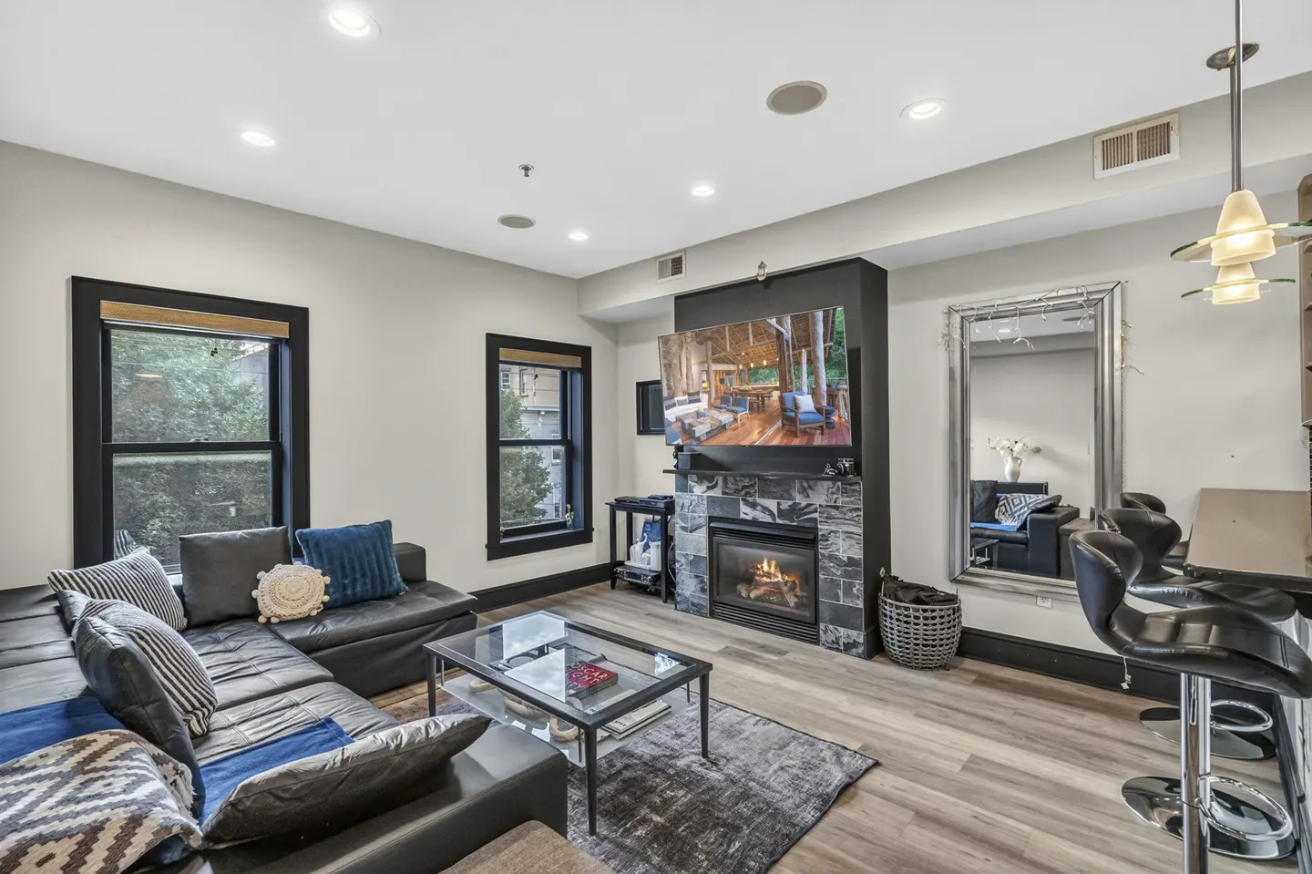 Modern living room with black leather sectional, glass coffee table, stone fireplace with TV, and a large wall mirror.