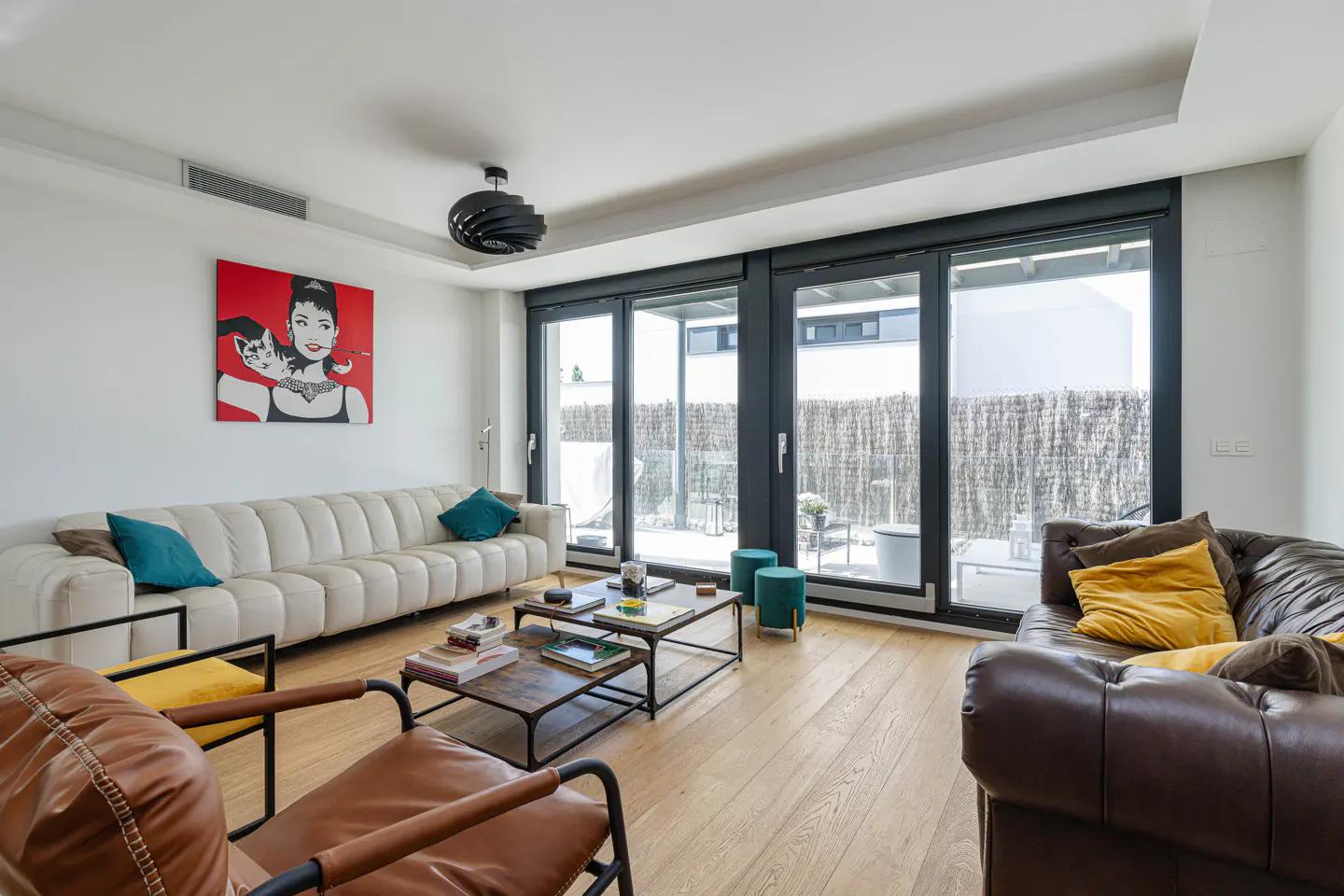 Living room with a white sofa, brown leather chairs, and a painting of Audrey Hepburn on the wall. Sliding glass doors lead to a patio.