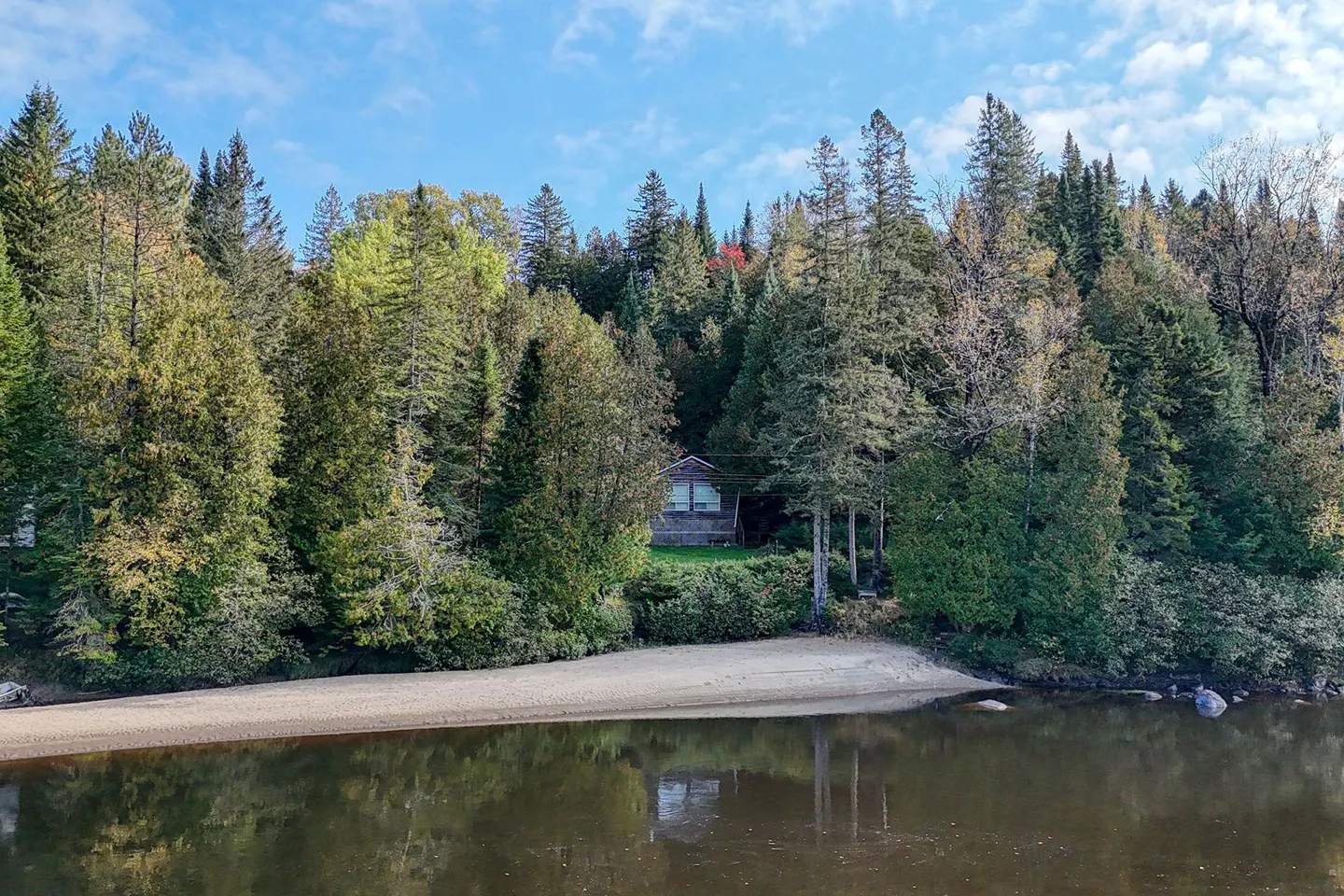 A small cabin nestled in a dense forest behind a sandy beach on a calm lake under a bright blue sky.