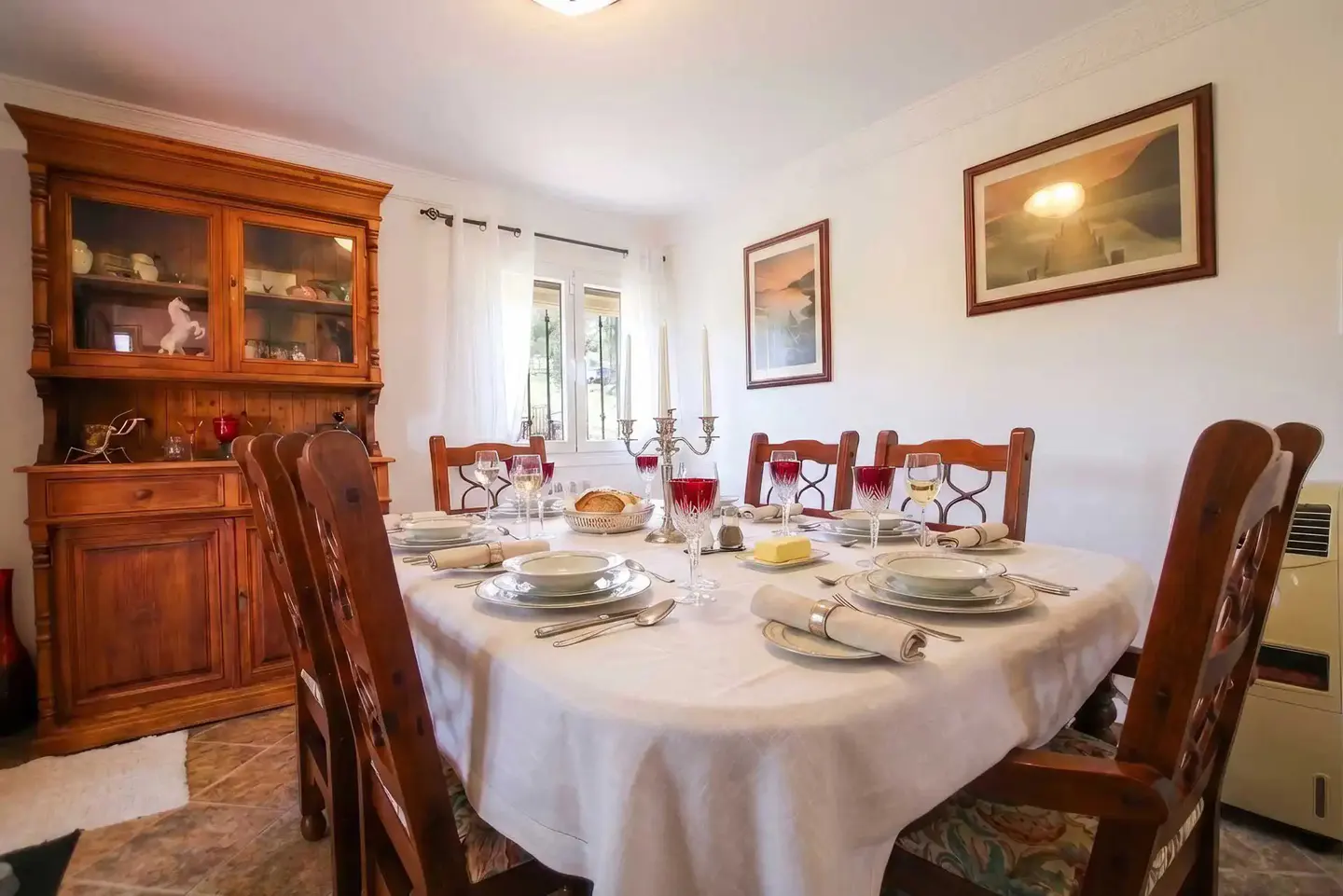 A dining room with a set table, wooden chairs, and a large wooden hutch. Two framed pictures hang on the white walls.