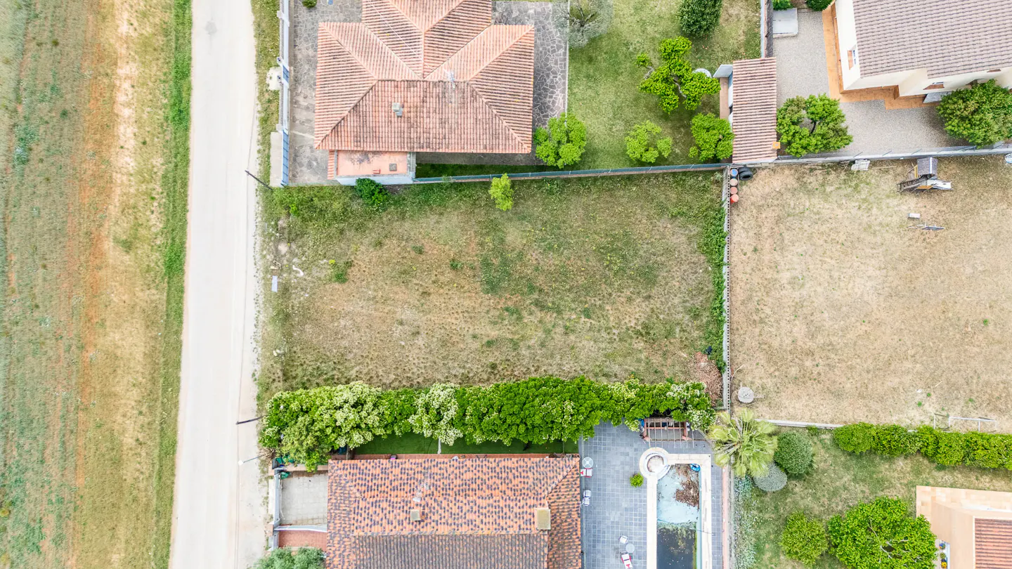 Aerial view of a vacant lot with dry grass, bordered by houses with red tile roofs, green trees, and a road.