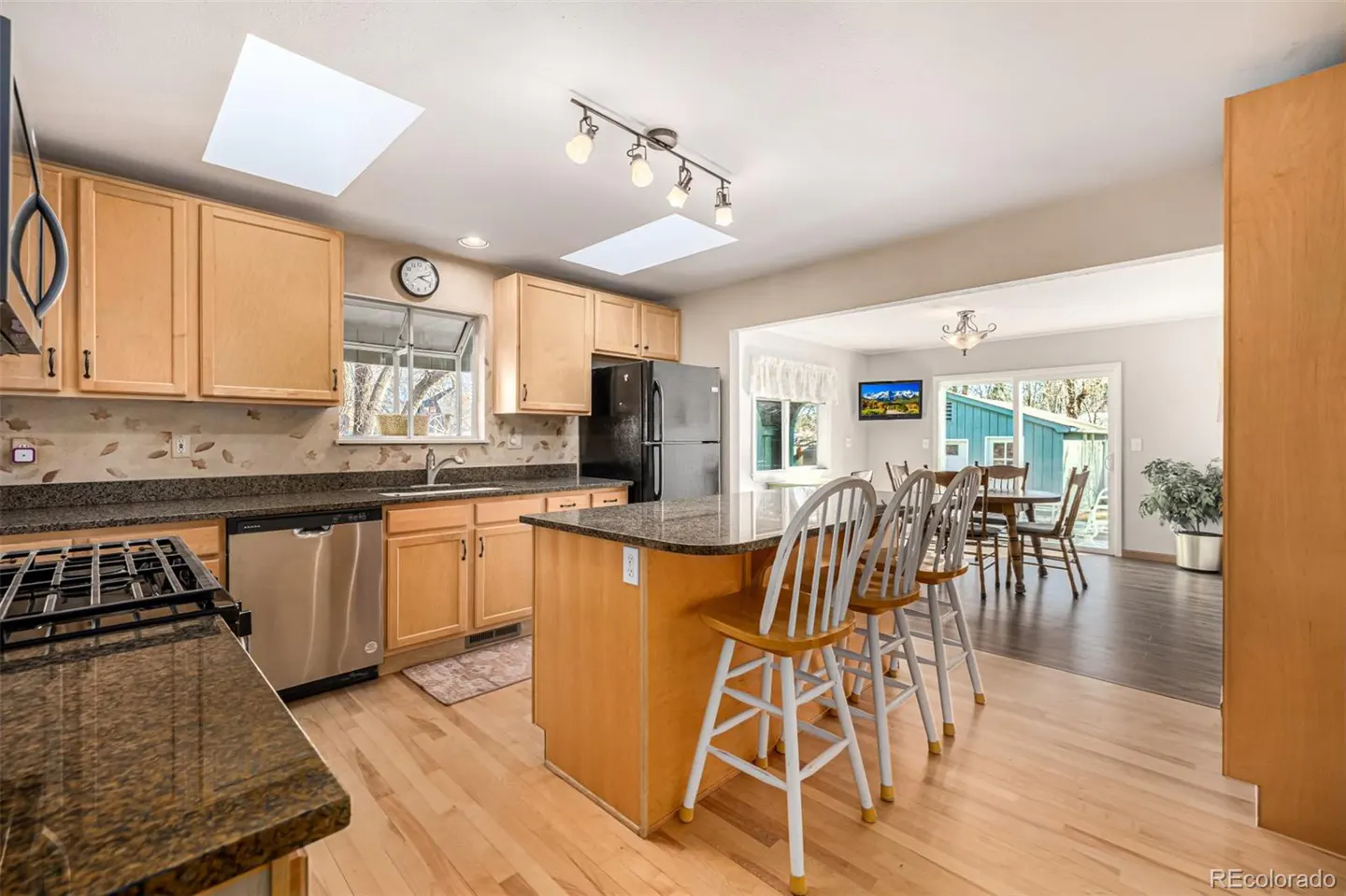 A bright kitchen with wood cabinets, granite countertops, and stainless steel appliances. A kitchen island with bar stools faces a dining area.