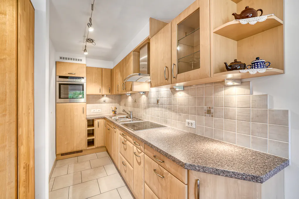 A well-lit kitchen with light wood cabinets, granite countertops, and tile backsplash. Teapots sit on shelves above the counter.