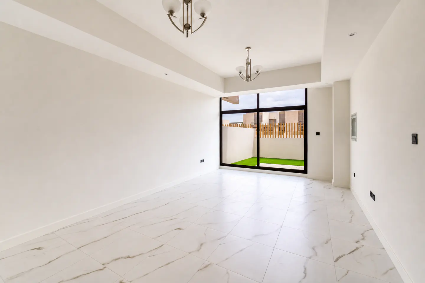 Bright, empty room with white walls, marble floors, and two chandeliers. A large window shows a fenced-in patio with artificial grass.