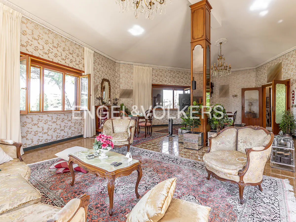 A spacious living room with patterned wallpaper, ornate furniture, and a large oriental rug. Natural light streams through the open windows.