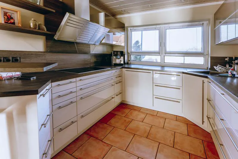 Bright kitchen with white cabinets, dark countertops, and terracotta tile flooring. Stainless steel range hood and large windows.
