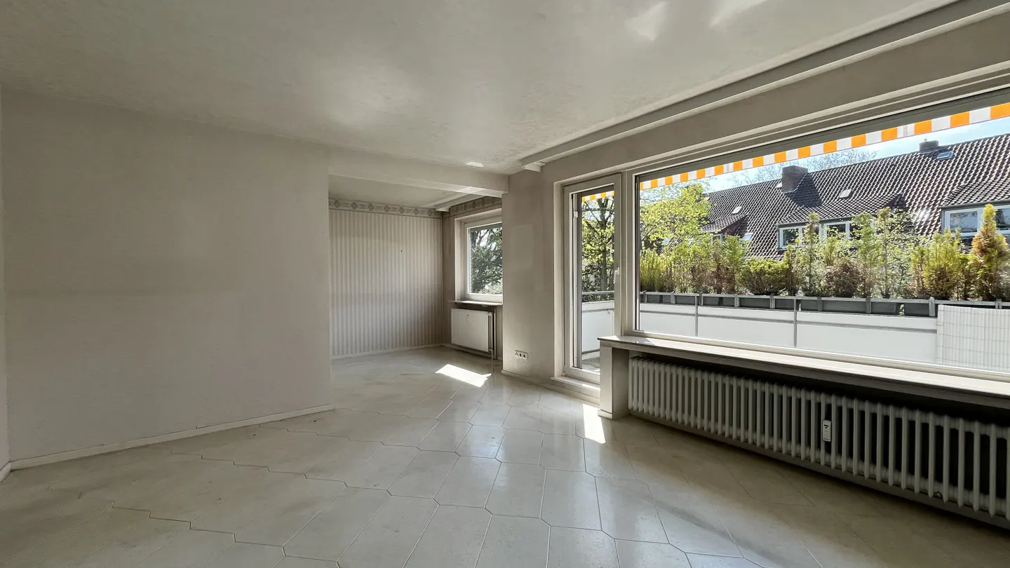 Bright, empty room with white walls, patterned floor tiles, and large windows overlooking a balcony with greenery.