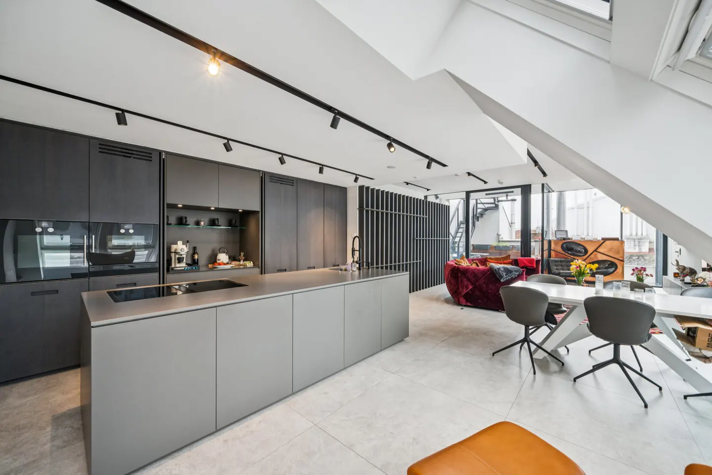 Open-concept kitchen and living area with gray cabinets, island, and dining table with gray chairs. A red velvet sofa is visible in the background.