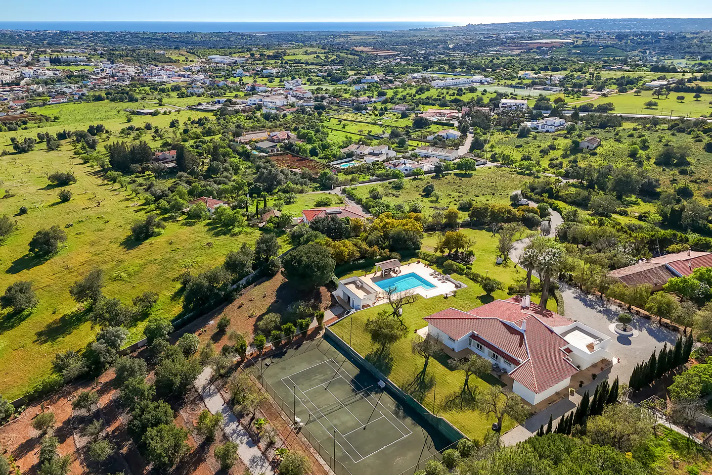 Aerial view of a large property with a tennis court, pool, and red-roofed house surrounded by green lawns and trees. A town is visible in the distance.
