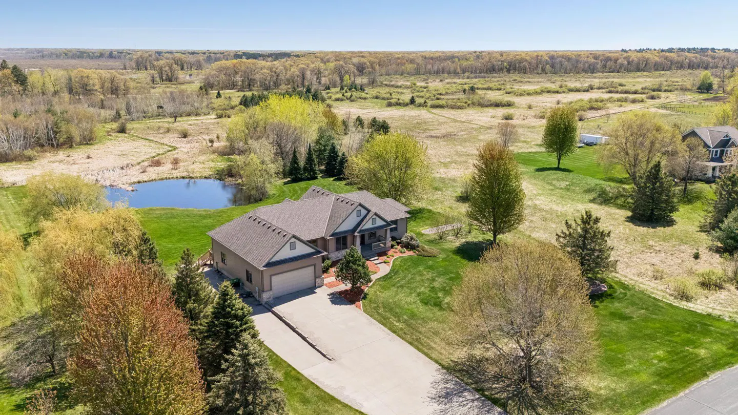 Aerial view of a tan house with a gray roof, a pond, and a long driveway surrounded by green grass and trees.