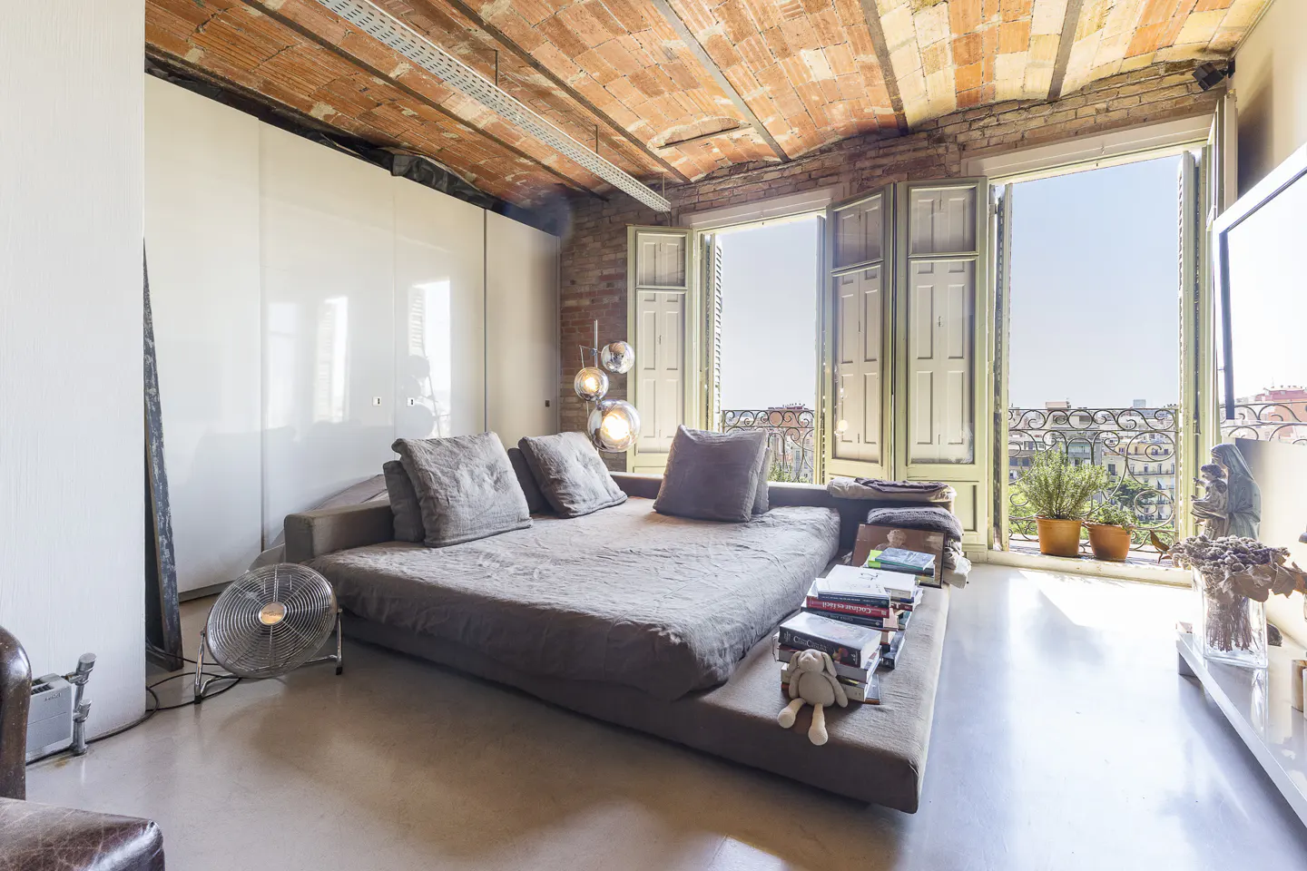 Loft apartment with a large platform bed, exposed brick ceiling, and open doors to a balcony with city views.