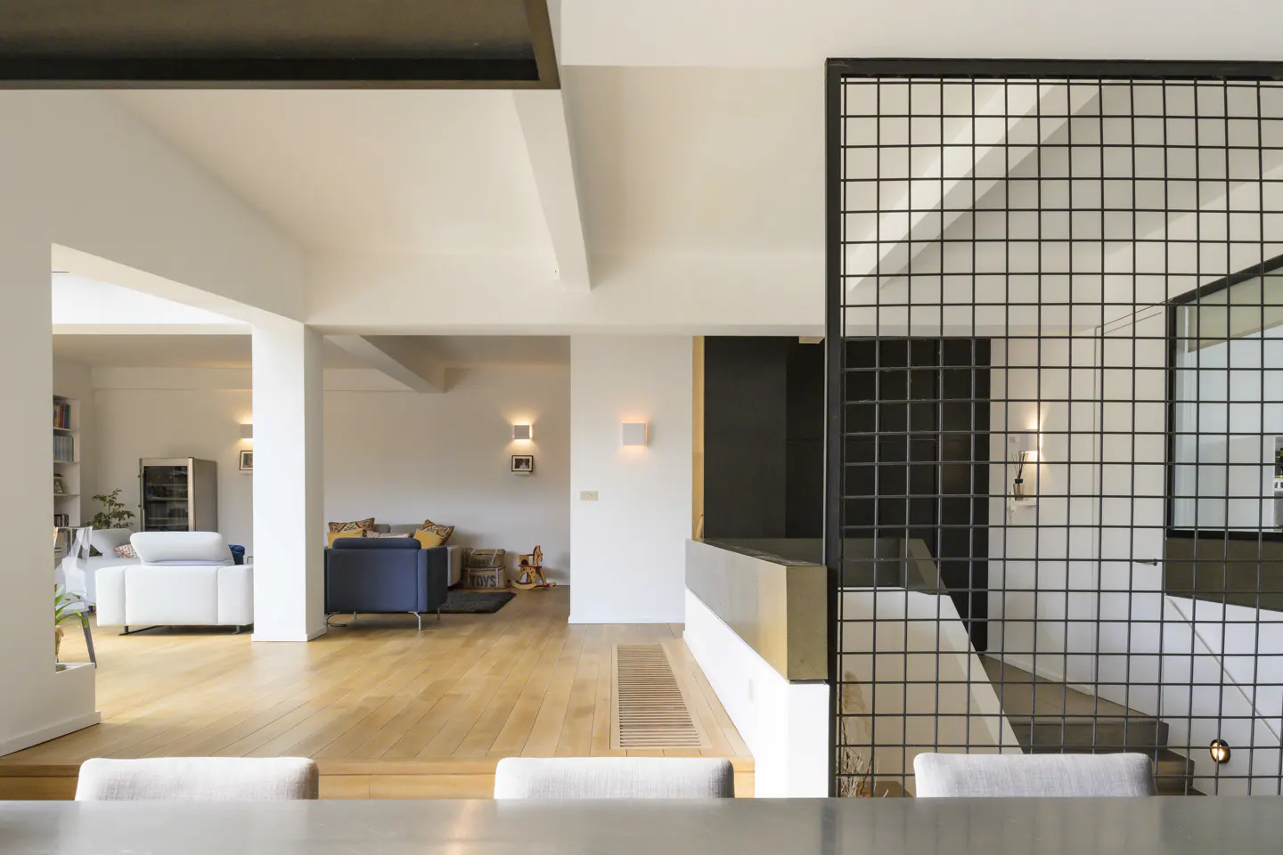 Open-concept living space with hardwood floors, white walls, and a black metal grid staircase. A blue sofa and white sofa are visible in the background.