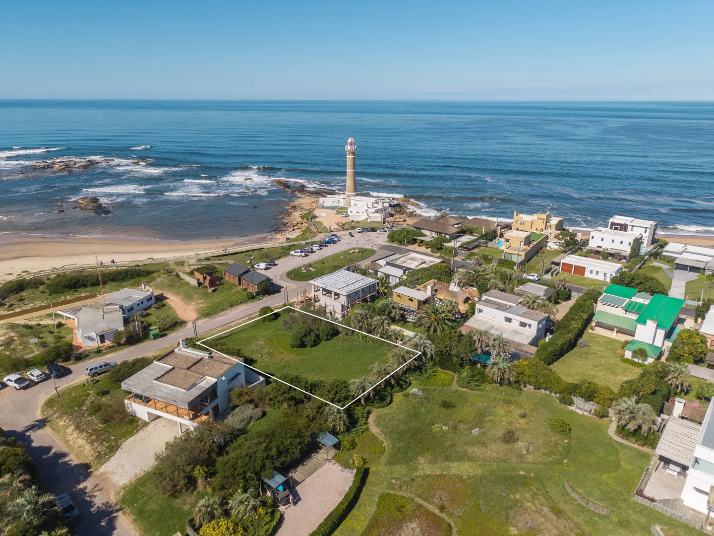 Aerial view of a grassy lot outlined in white, near a beach, lighthouse, and houses under a clear blue sky.