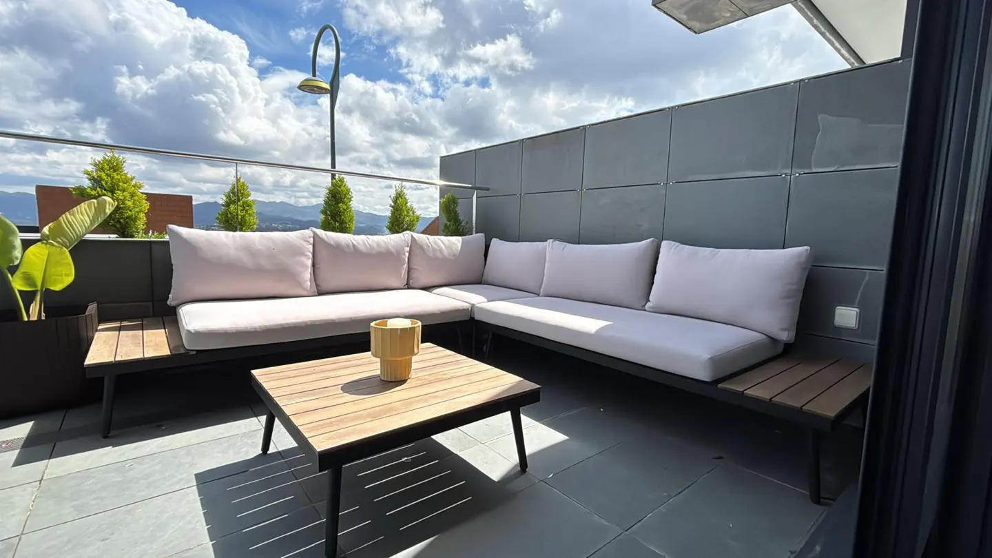 Modern balcony with L-shaped light gray sofa, wooden coffee table, and potted plant against a backdrop of distant mountains.