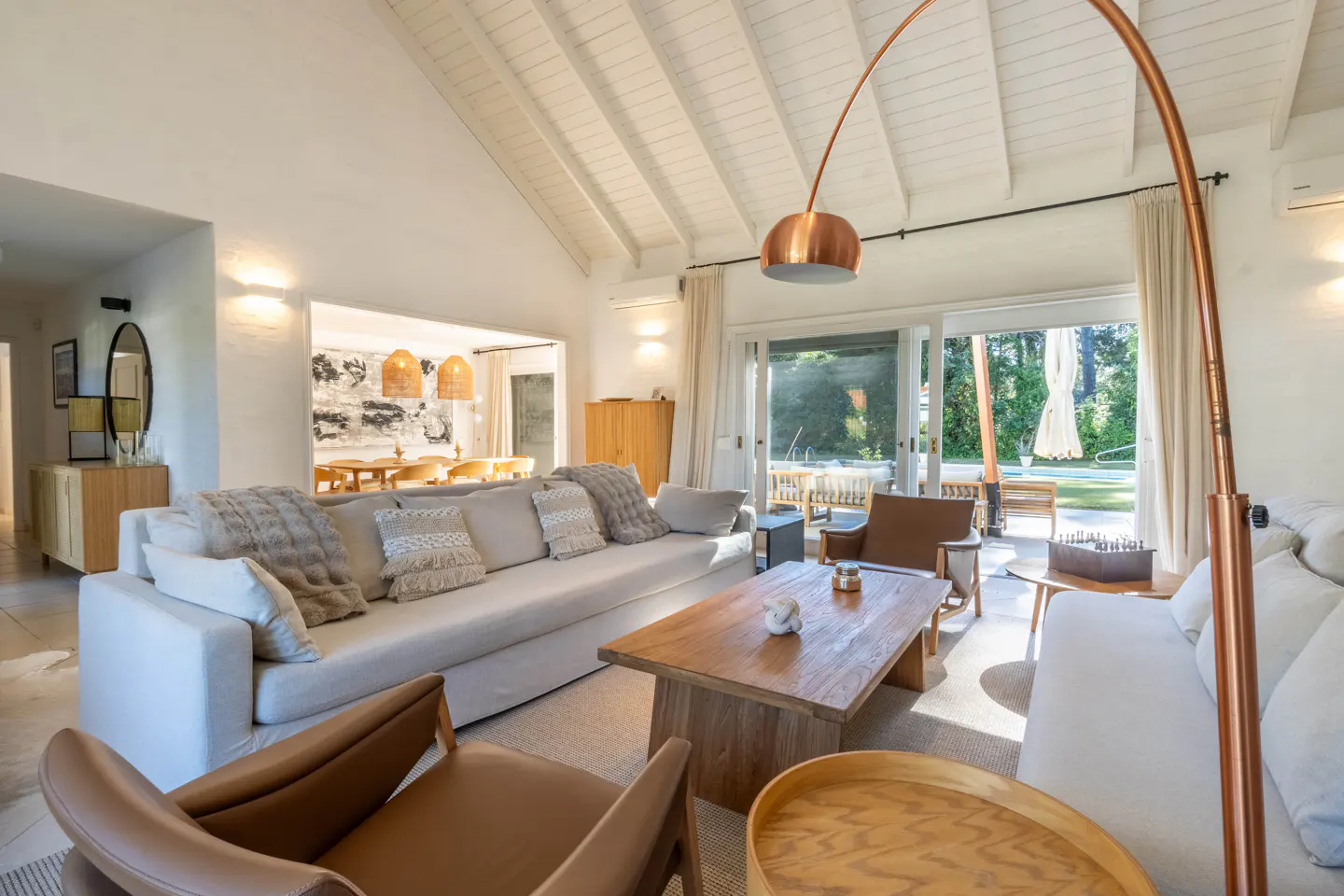 Bright living room with white walls, vaulted ceiling, and large windows. Sofas, wood coffee table, and copper floor lamp furnish the space.