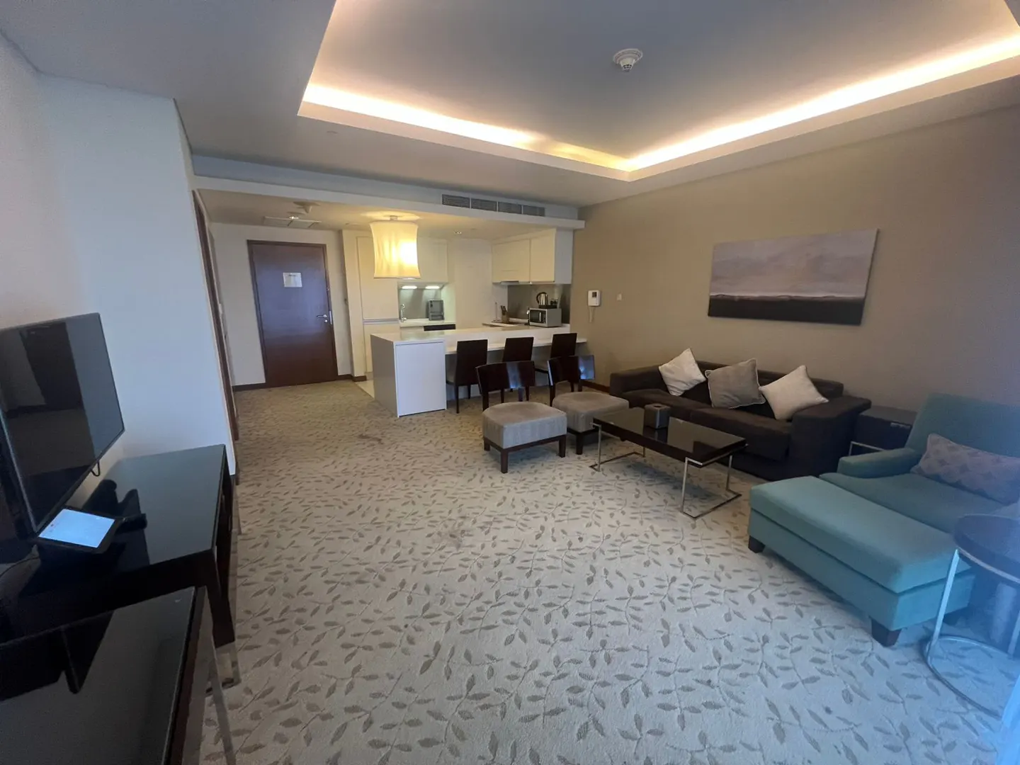 A wide shot of a hotel suite with a kitchen, living room, and a leaf-patterned carpet. The living room has a brown sofa and a blue chaise lounge.