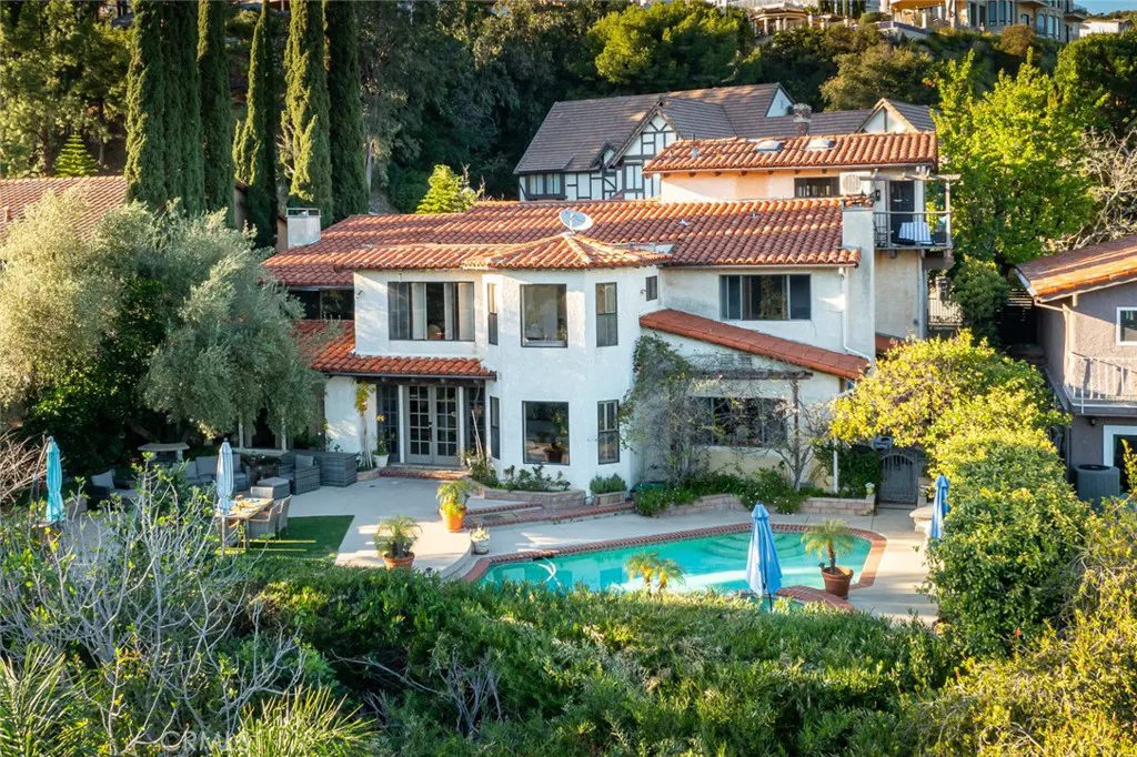 Two-story white Mediterranean home with a red tile roof, swimming pool, and patio surrounded by lush trees and greenery.