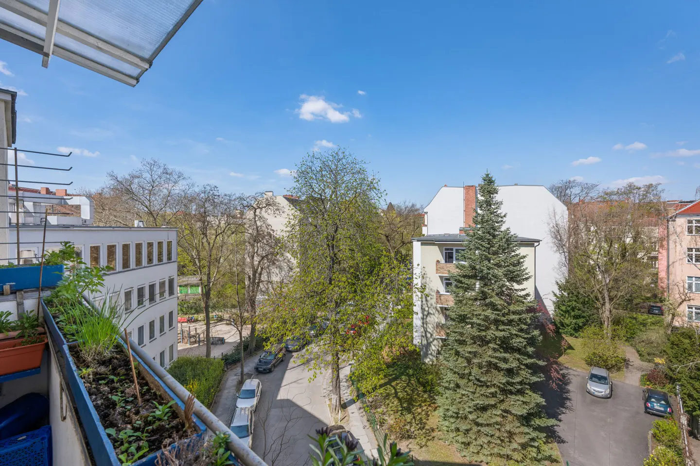 View from a balcony overlooking a quiet street with green trees, parked cars, and surrounding residential apartment buildings.