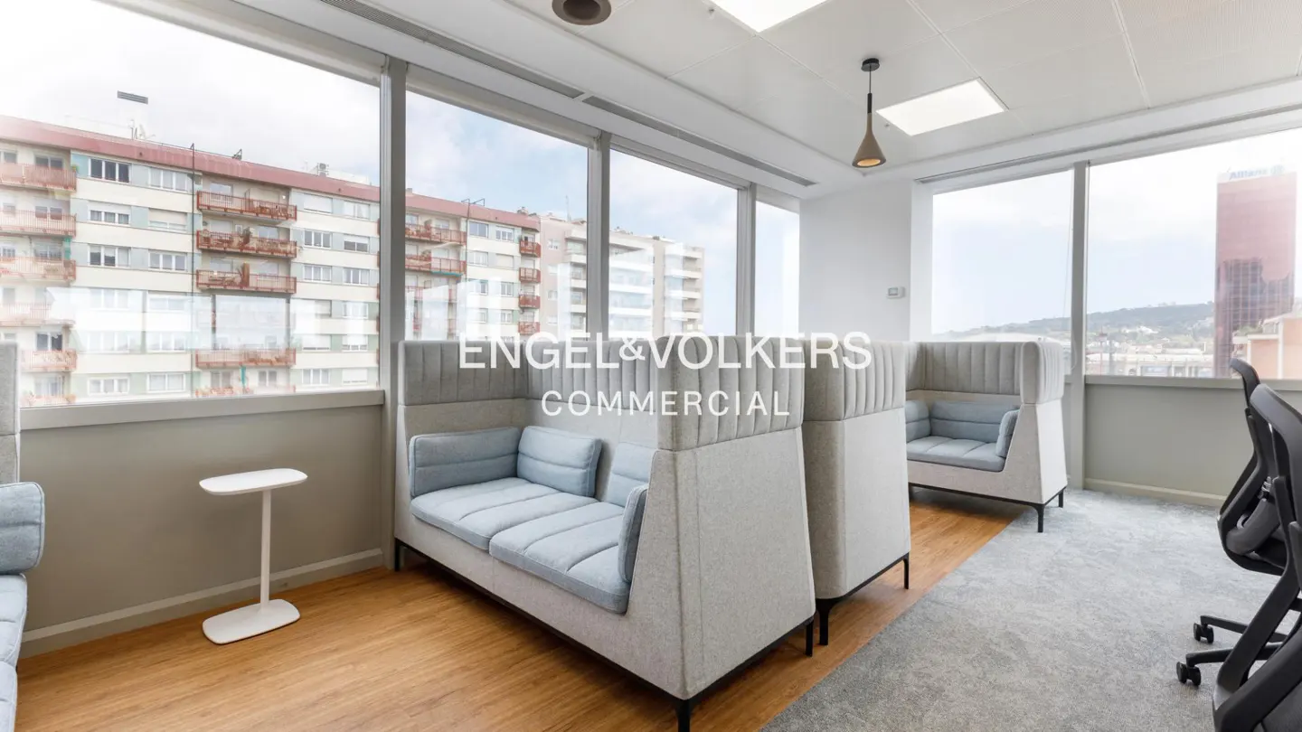 Bright office space with light gray sofas, tall windows showing city views, and Engel & Volkers Commercial logo.