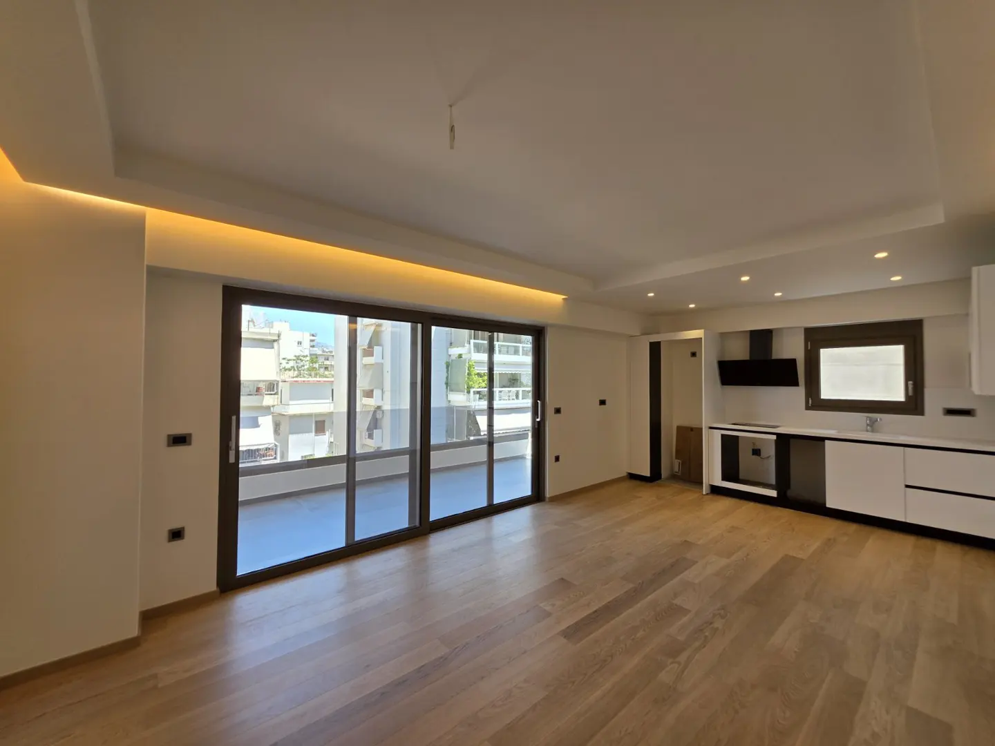 Modern open-plan living room with hardwood floors, recessed lighting, and a sleek white kitchen with a balcony view.
