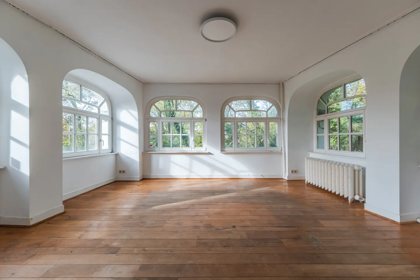 Bright, empty room with hardwood floors, white walls, and arched windows showing trees outside. A white radiator sits under one window.