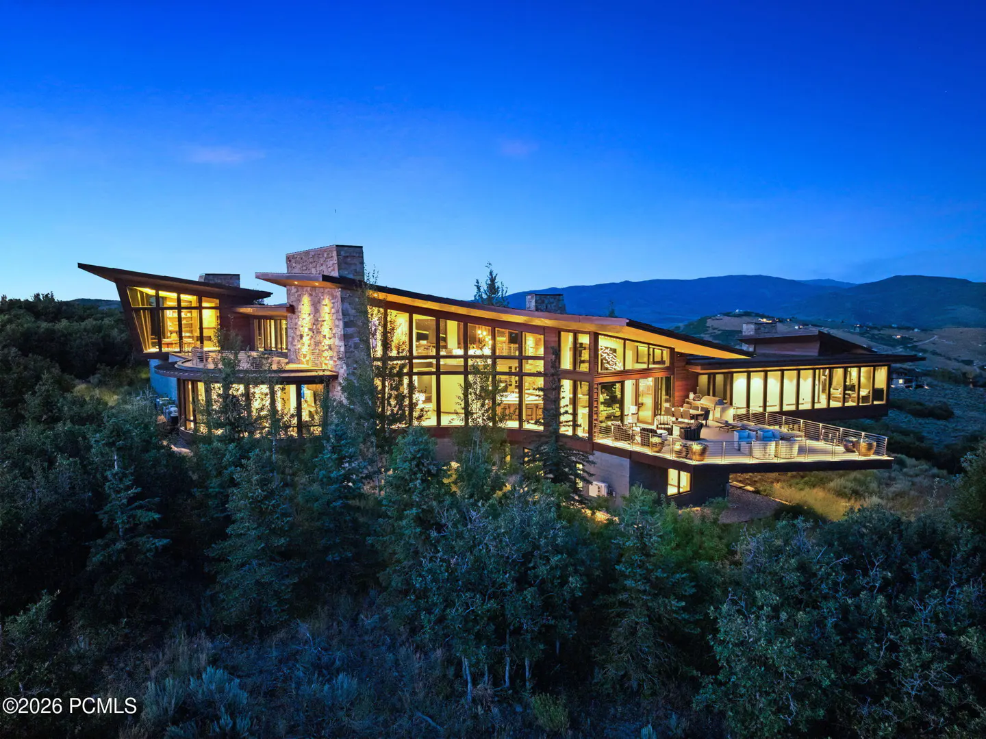 Modern luxury home with stone accents and large windows, nestled in a wooded area under a blue twilight sky.
