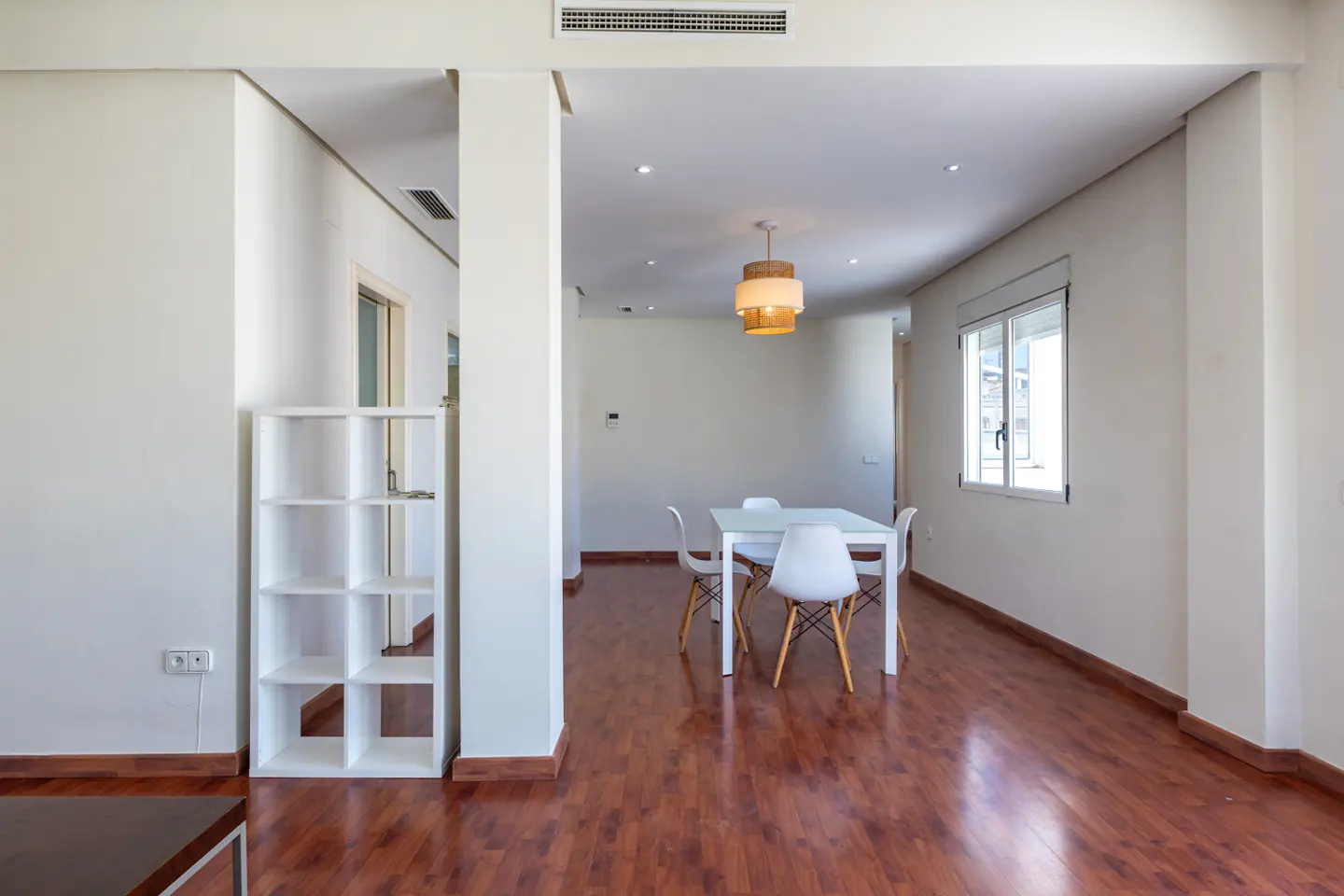 Bright, open-plan dining area with white walls, wood floors, and a white table with four chairs. A modern light fixture hangs above.