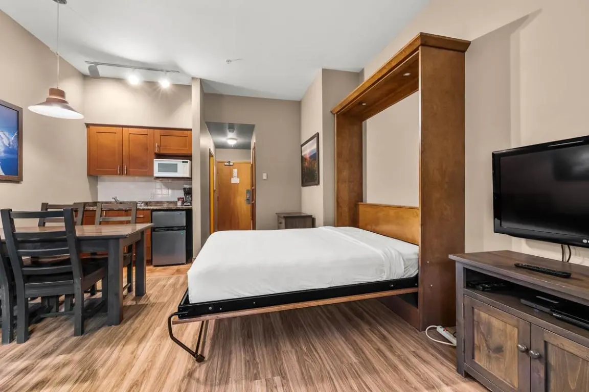 Studio apartment with a Murphy bed, kitchenette, dining table, and TV on a wooden stand.