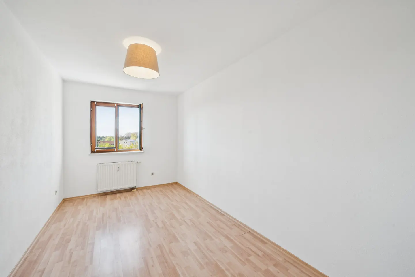 Bright, empty room with wood floors, white walls, and a window with a view. A tan lampshade hangs from the ceiling.