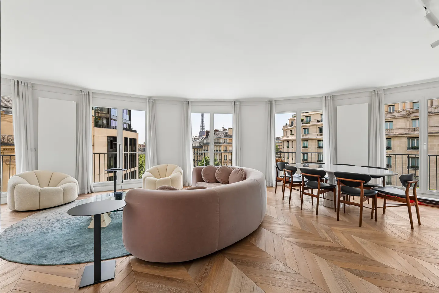 Modern living room with a curved pink sofa, cream chairs, dining set, and a view of the Eiffel Tower through large windows.