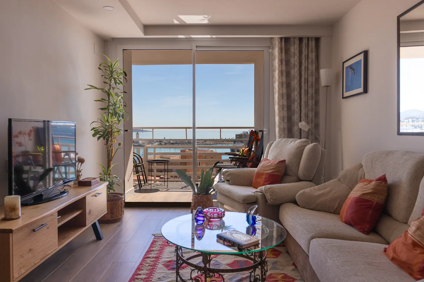 Living room with a beige sofa, glass table, and TV. Balcony with ocean view in the background.