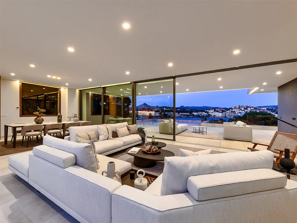 Modern open-plan living room with white sofas, dining area, and floor-to-ceiling glass doors overlooking a coastal sunset.