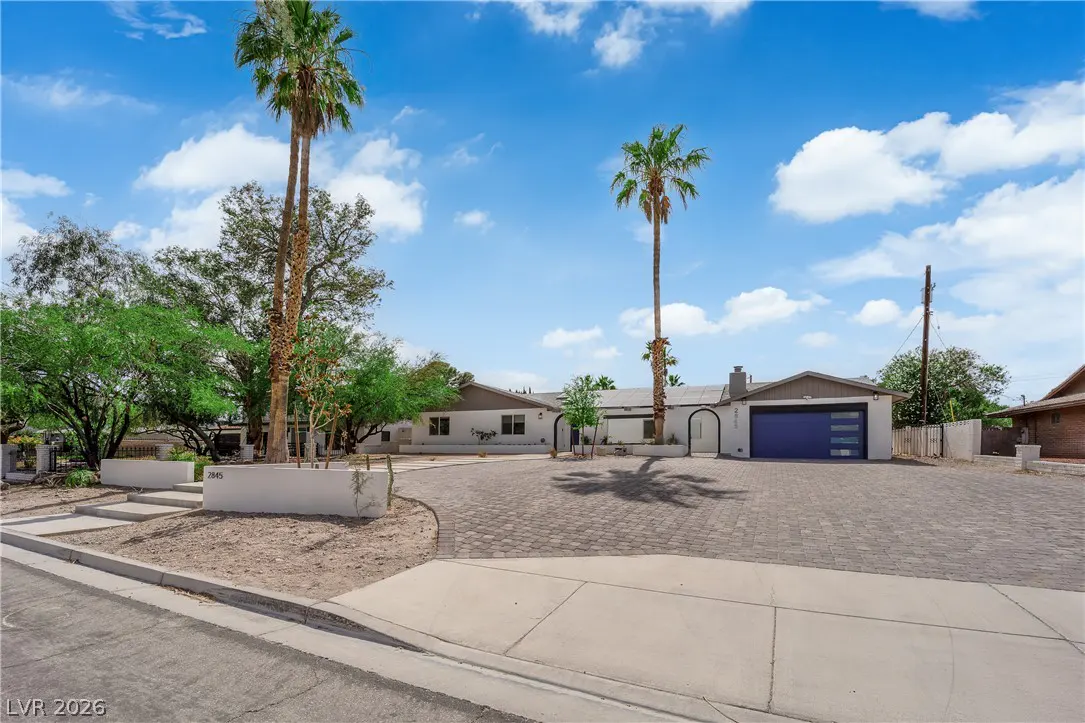 Single-story white home with a blue garage door, large paved driveway, and tall palm trees under a blue sky.