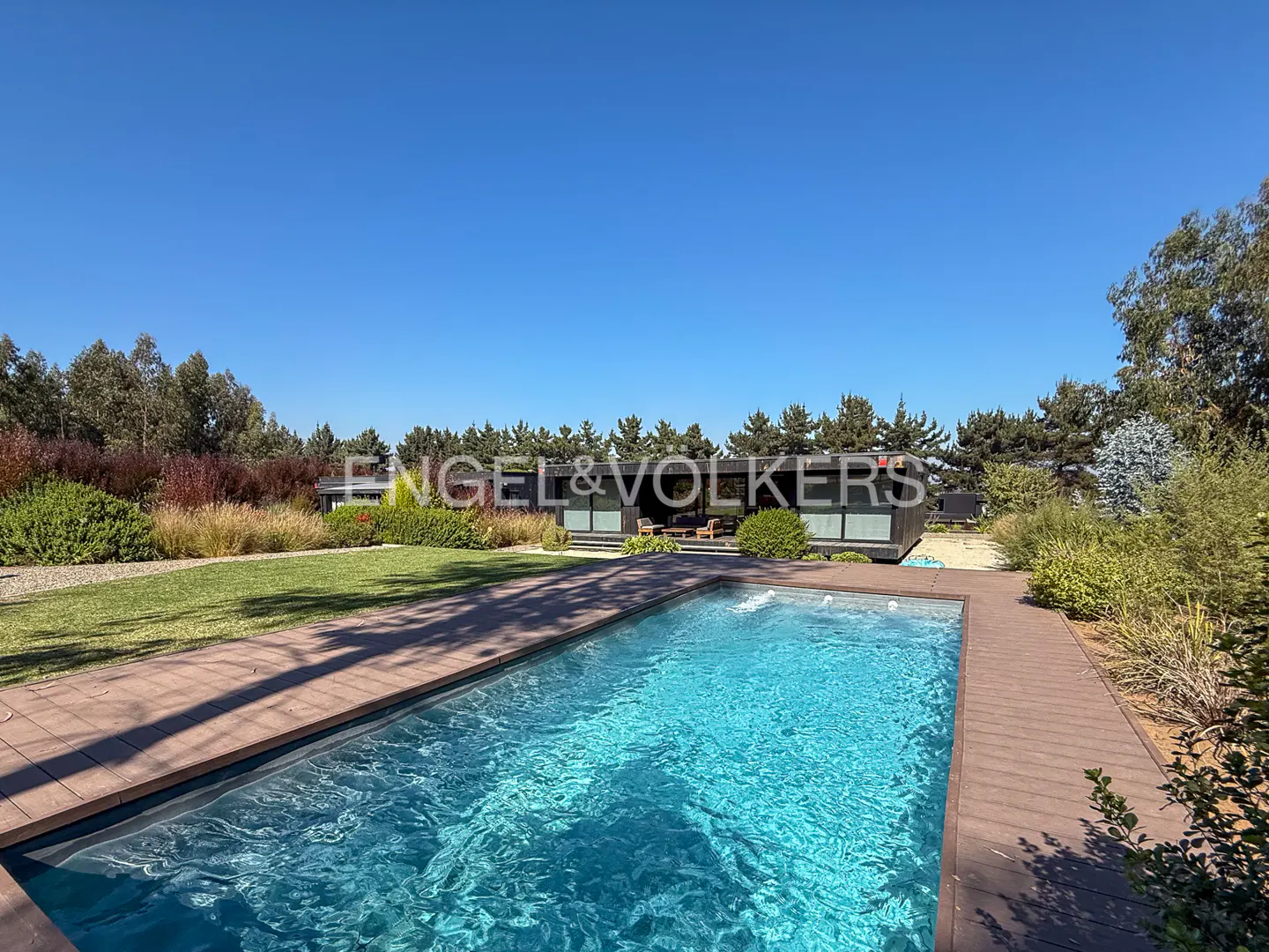 A wide shot of a modern home with a pool. The pool is turquoise and surrounded by a brown deck. The house is dark gray with large windows. The sky is blue.