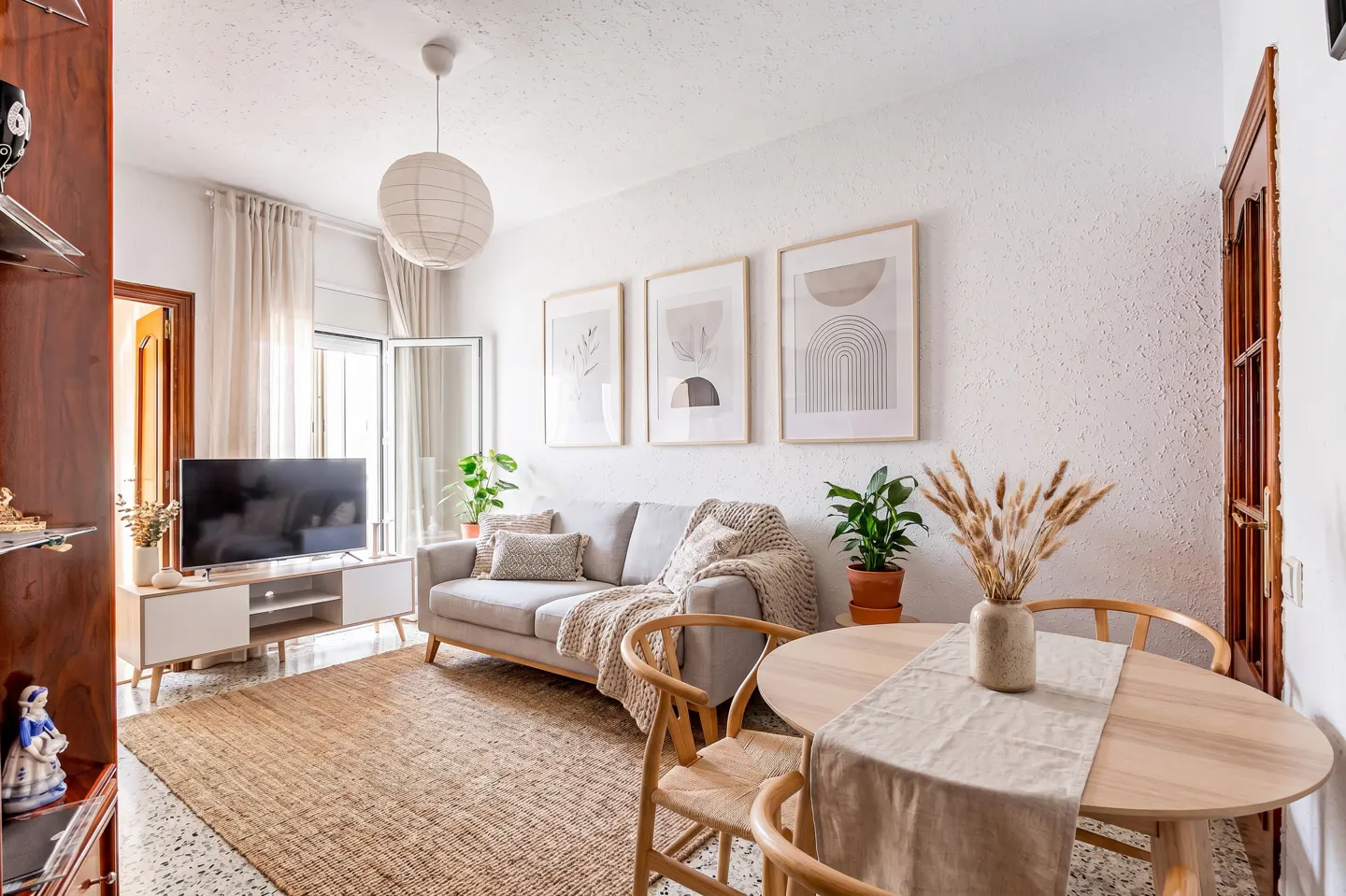 Bright living room with a gray sofa, round wooden table, and three framed art prints on a white wall. A TV sits on a white stand.