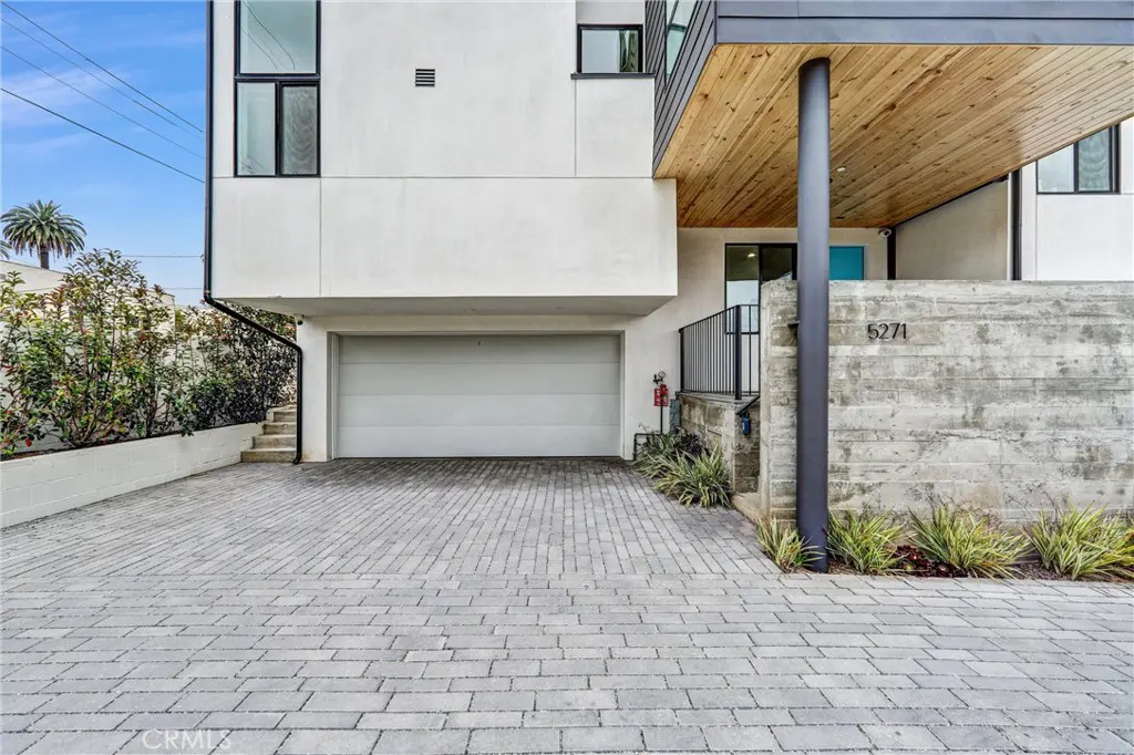 Modern white house with a gray brick driveway, a gray garage door, and a wooden porch ceiling. The house number 5271 is visible on a concrete wall.