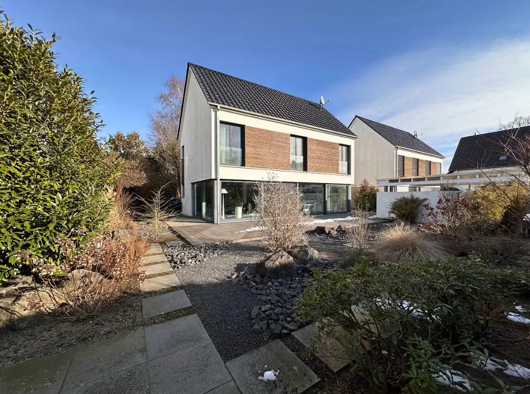 Modern two-story house with white siding, dark roof, and wood accents. Landscaped yard with stone path and greenery.