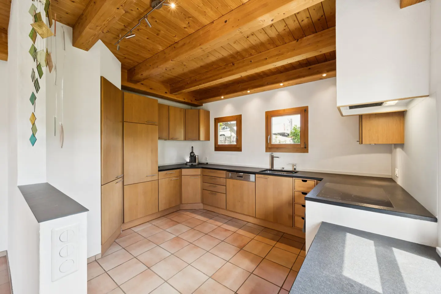 A bright kitchen with light wood cabinets, dark countertops, and a wood-beamed ceiling. Two windows let in natural light.