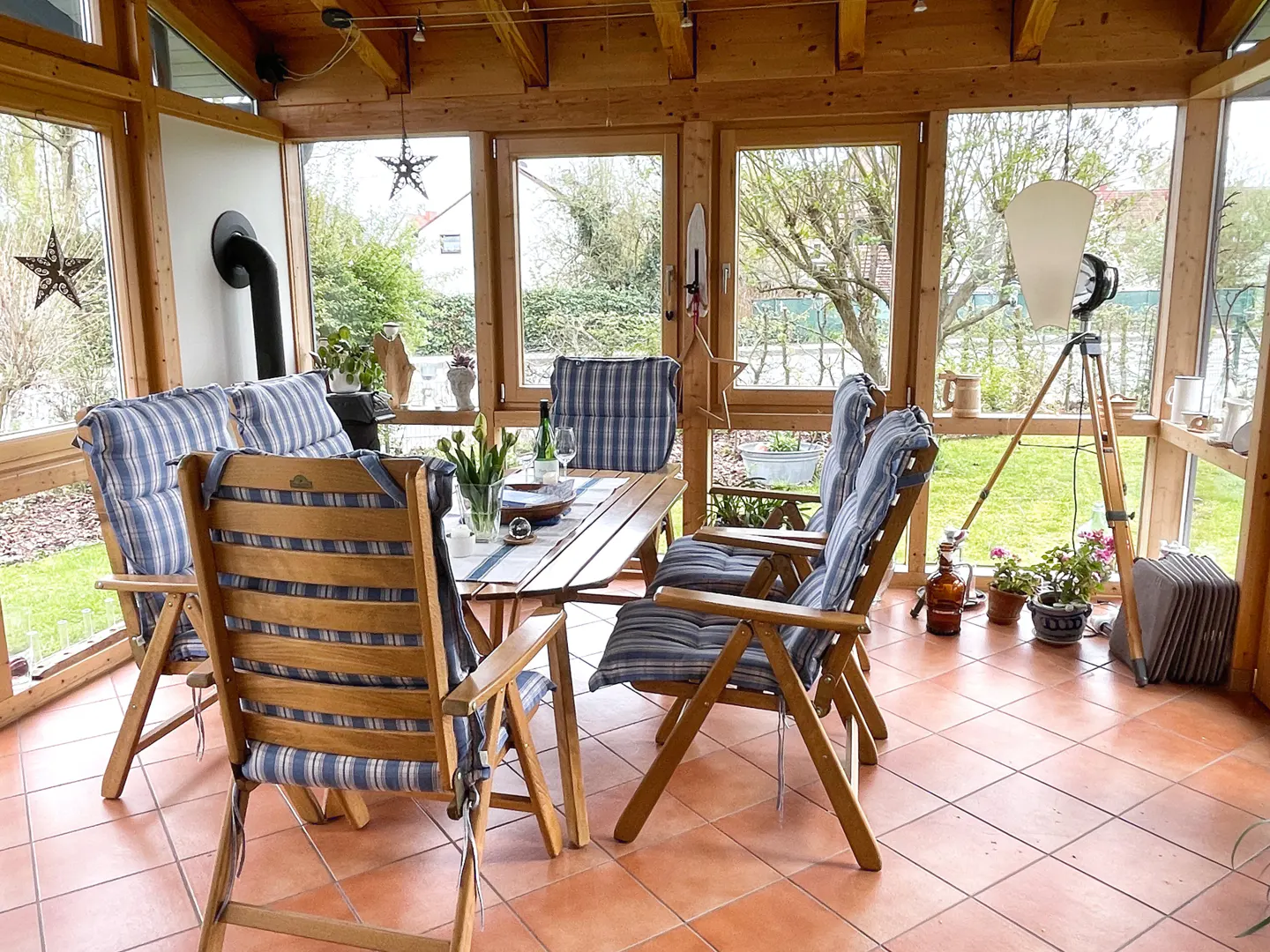 Sunroom with wood-framed windows, terracotta tile floor, and a wood table surrounded by chairs with blue striped cushions.