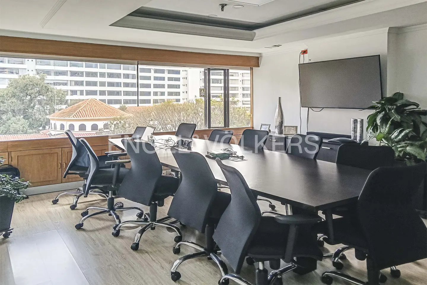 Conference room with a long dark table, black chairs, and a large window with a city view. A TV is mounted on the wall.