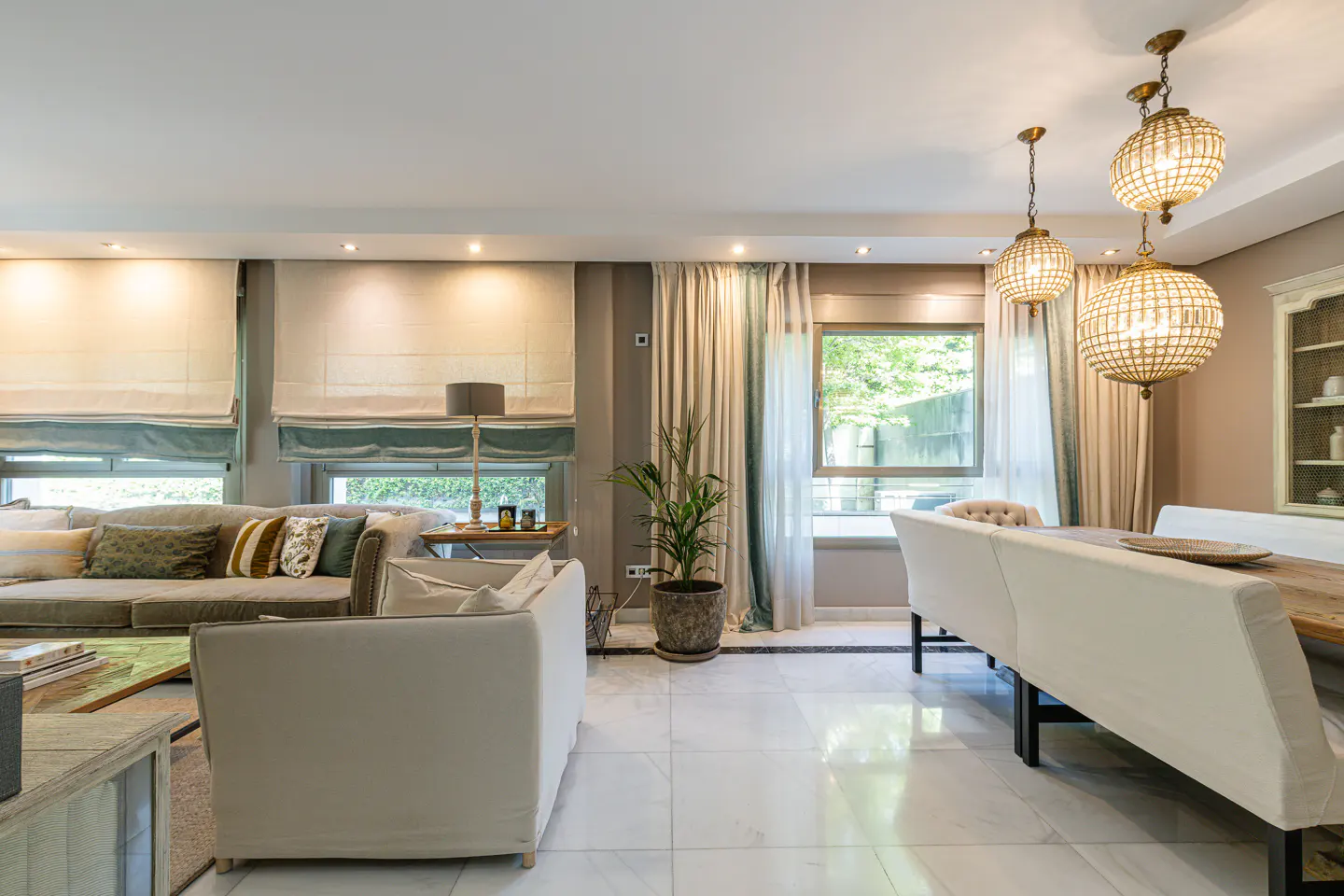 Bright, open-plan living and dining area with marble floors. A large sofa, dining table with white chairs, and crystal chandeliers are visible.