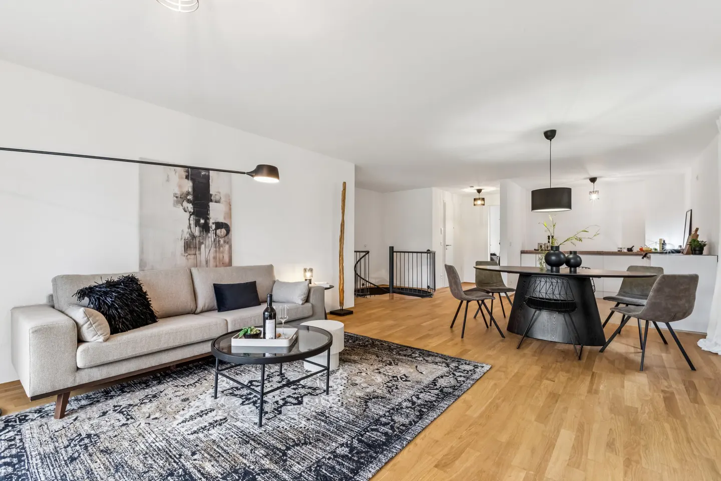 Bright, open-concept living space with a beige sofa, black pillows, and a patterned rug. A round dining table with chairs sits on hardwood floors.