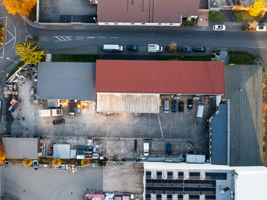 Aerial view of commercial buildings with red and gray roofs, parking lots, and solar panels. Cars and trucks are parked nearby.