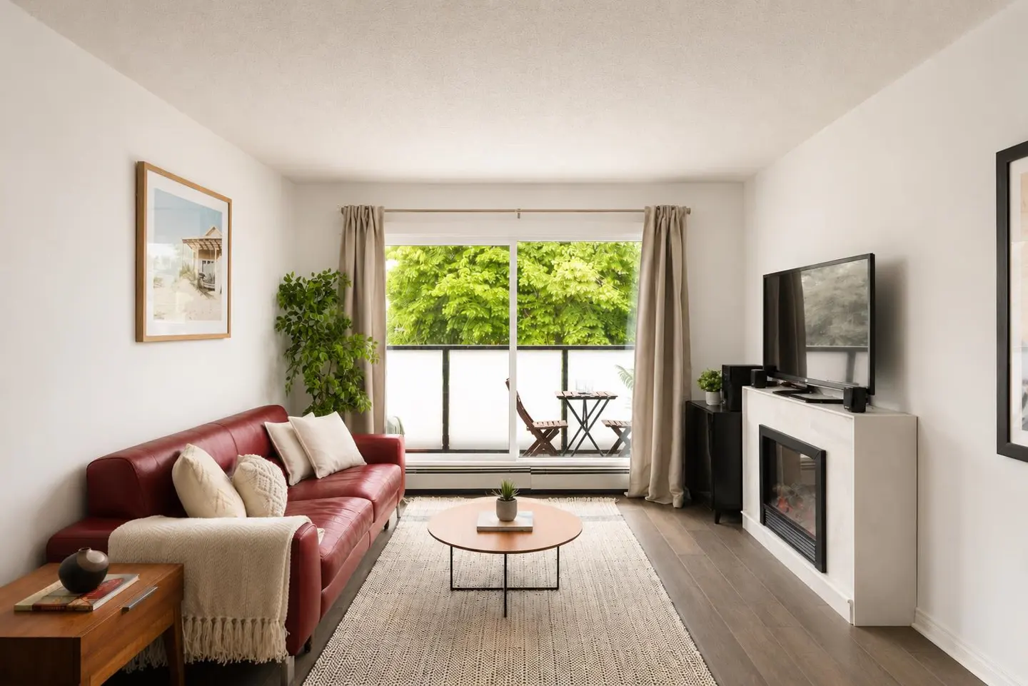 Bright living room with a red leather sofa, round coffee table, and a fireplace under a TV. Balcony with green trees visible.