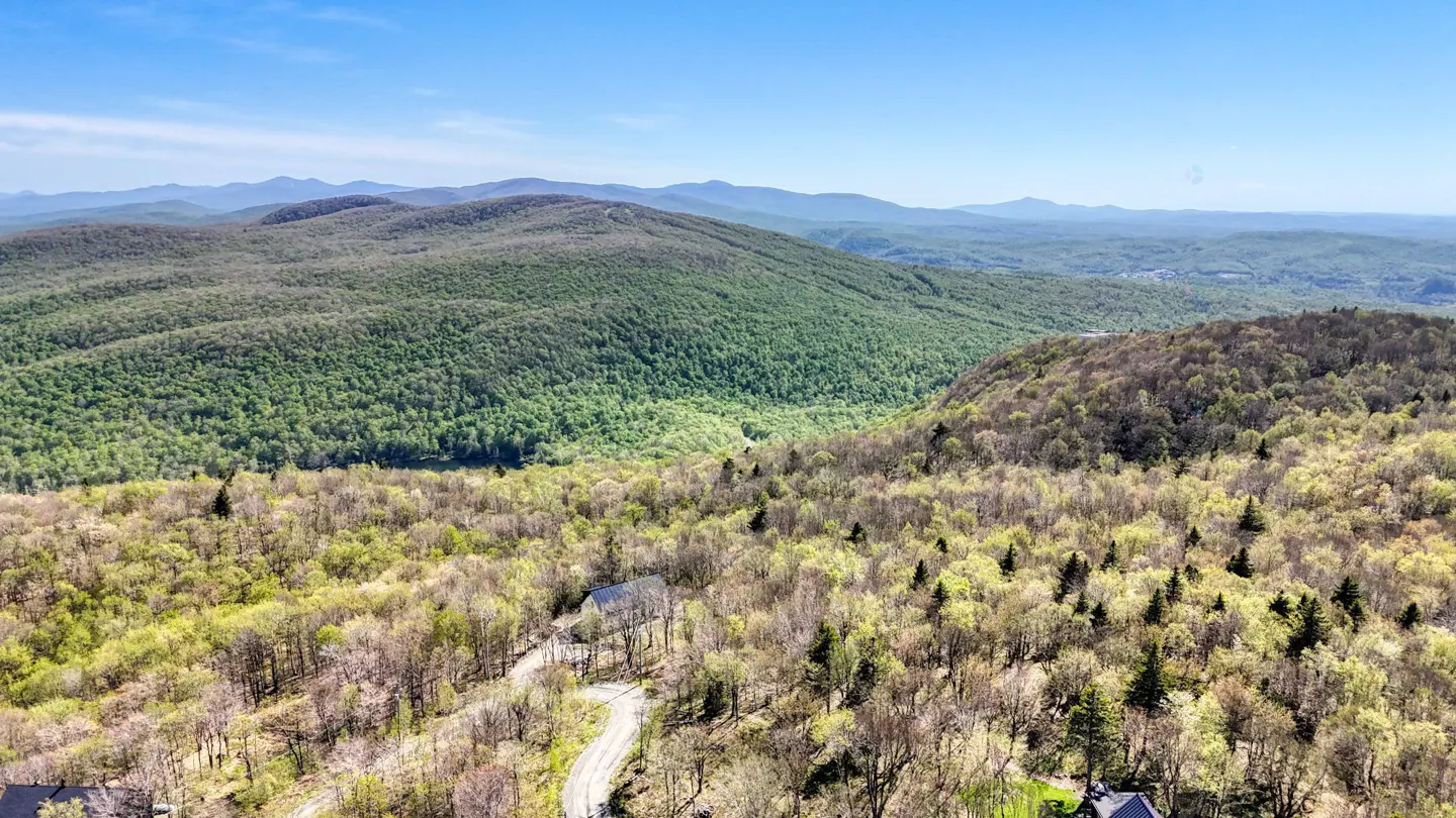 Aerial view of a house nestled in a lush, green mountain forest under a clear blue sky with distant mountain ranges.