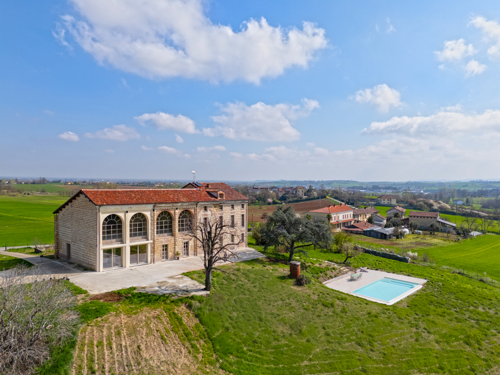 Aerial view of a stone house with a red tile roof, a pool, and green fields under a blue sky.