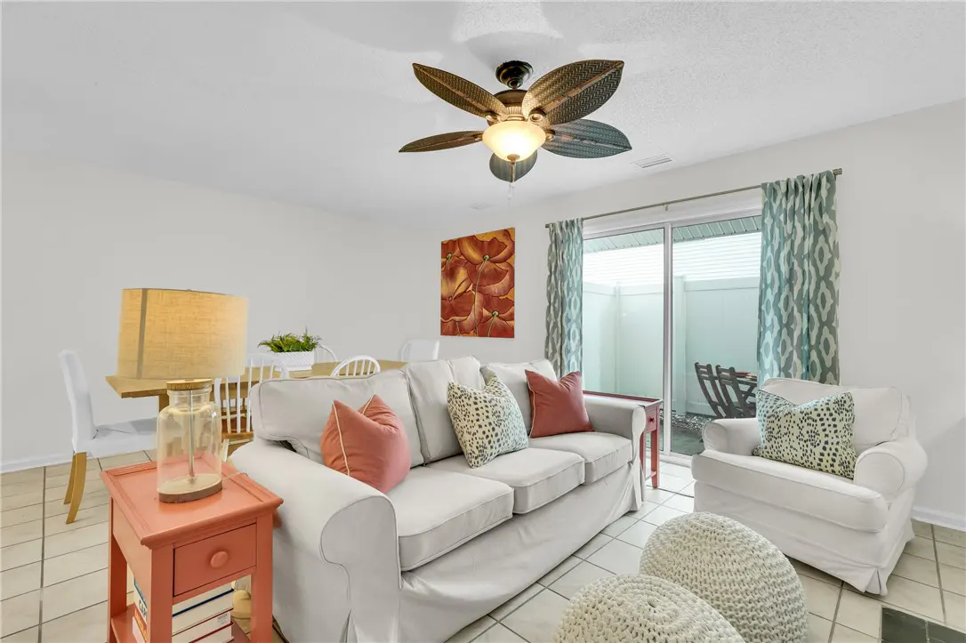 Bright living room with white sofa, chair, and poufs. A dining table and coral side table add color. Sliding glass doors lead to a patio.