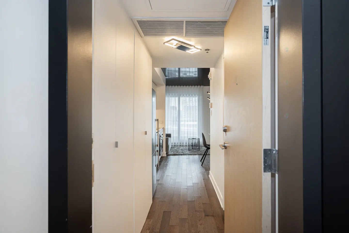 A bright condo hallway with white walls, wood floors, and a modern light fixture. An open door reveals a living area with a window and sheer curtains.