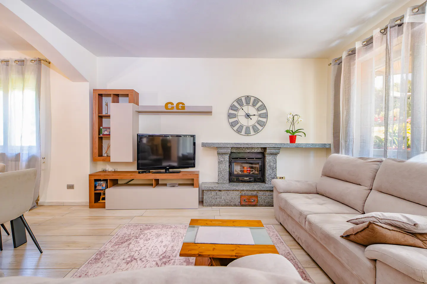 Bright living room with a beige sofa, wooden coffee table, stone fireplace, wall-mounted TV, and a large round wall clock.