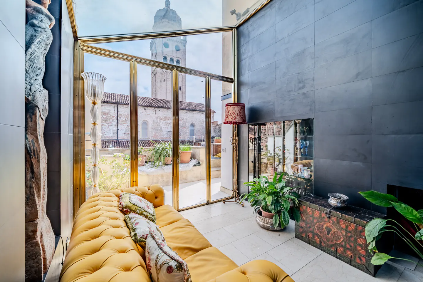 A bright living room with a yellow sofa, plants, and a view of a church tower through gold-framed glass doors.